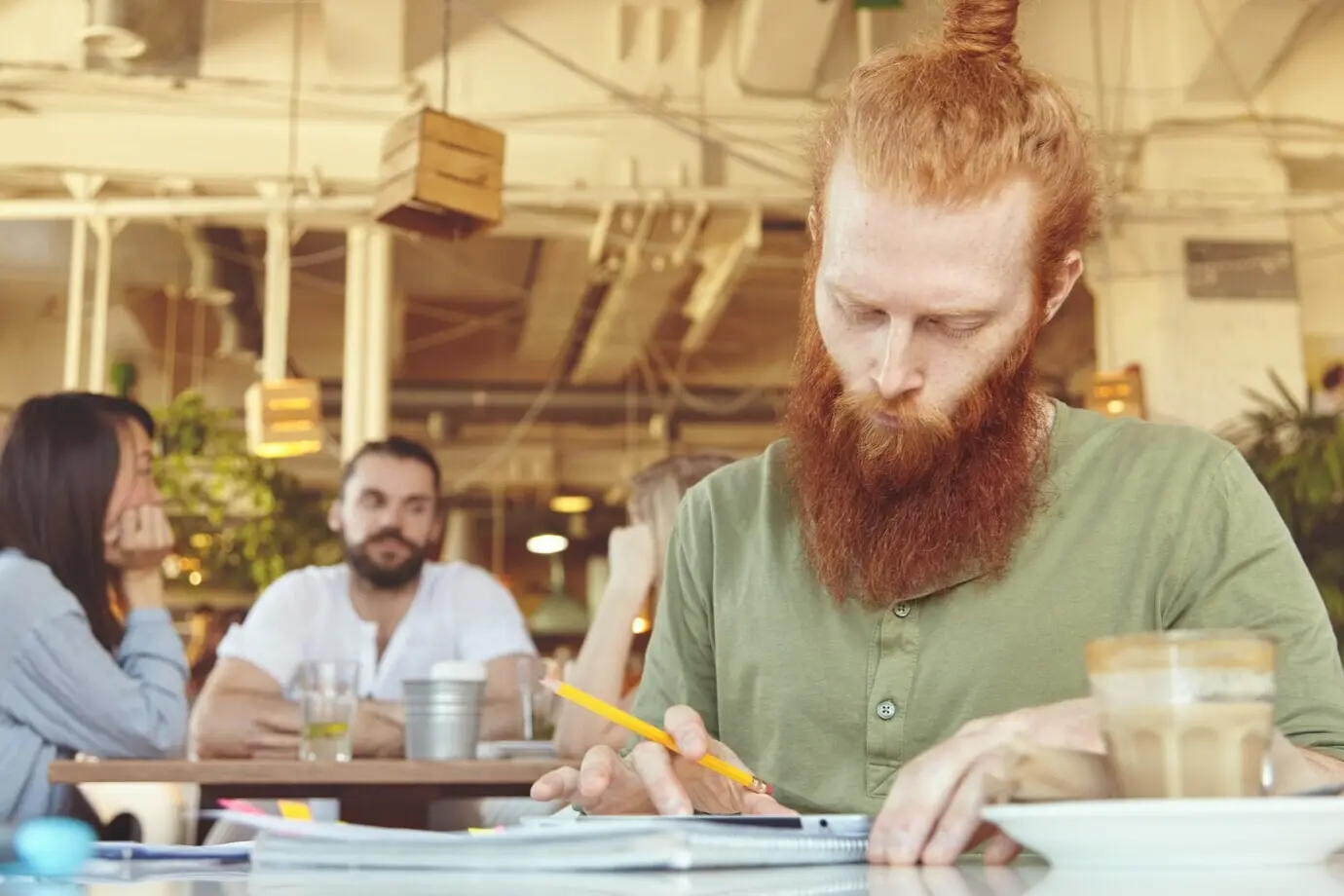 Freelancer hipster sosteniendo un lápiz, tomando notas en hojas de papel con gráficos y usando una tableta digital para trabajo a distancia en un espacio de coworking.